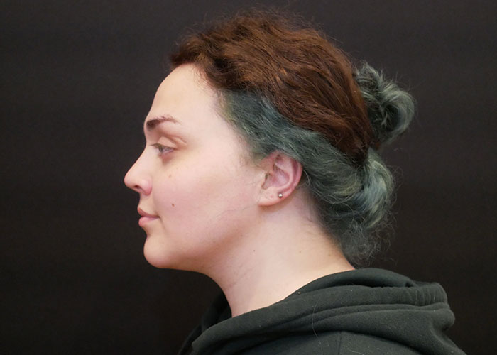 Profile of a woman with brown and teal hair in a bun, wearing a black hoodie against a dark background. Saxon MD