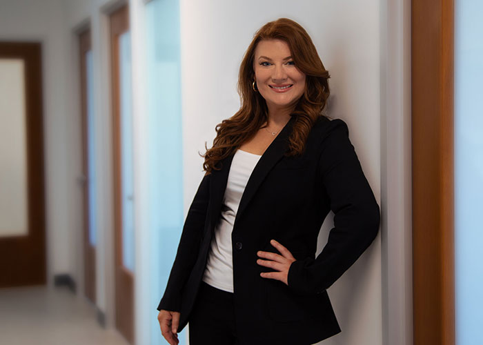 A confident woman with red hair wearing a black blazer smiles while standing in an office hallway. Saxon MD