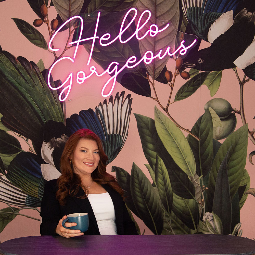 A woman smiling at the camera, holding a blue mug, seated in front of a floral wall with a neon sign reading "hello gorgeous. Saxon MD