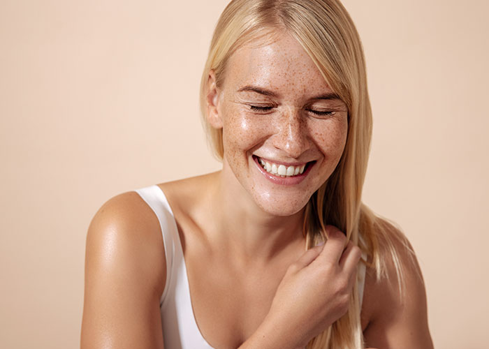 A joyful young woman with freckles and blonde hair, smiling brightly and wearing a white tank top, on a light peach background. Saxon MD