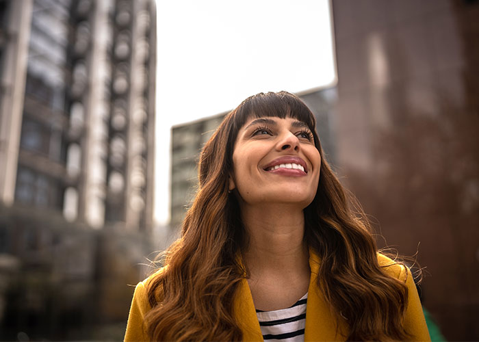 A smiling woman in a yellow coat looking upwards in an urban setting with high-rise buildings in the background. Saxon MD