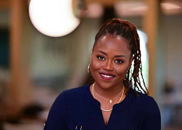 A smiling woman with braided hair, wearing a navy top and gold earrings, sitting in an office environment with soft-focus background lighting. Saxon MD