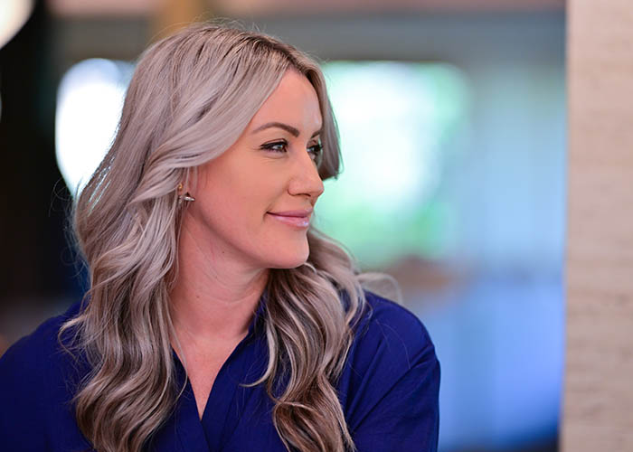 Profile view of a smiling woman with long gray hair, wearing a blue shirt, indoors with a blurred background. Saxon MD