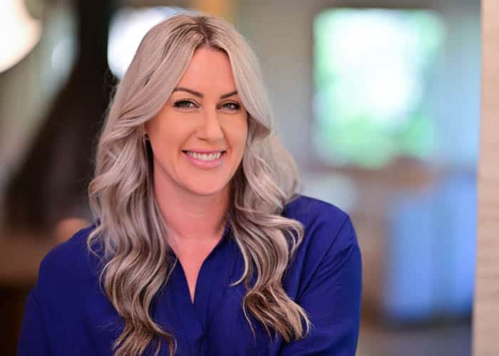 A smiling woman with long wavy gray hair, wearing a blue blouse, posing in a softly blurred indoor background. Saxon MD