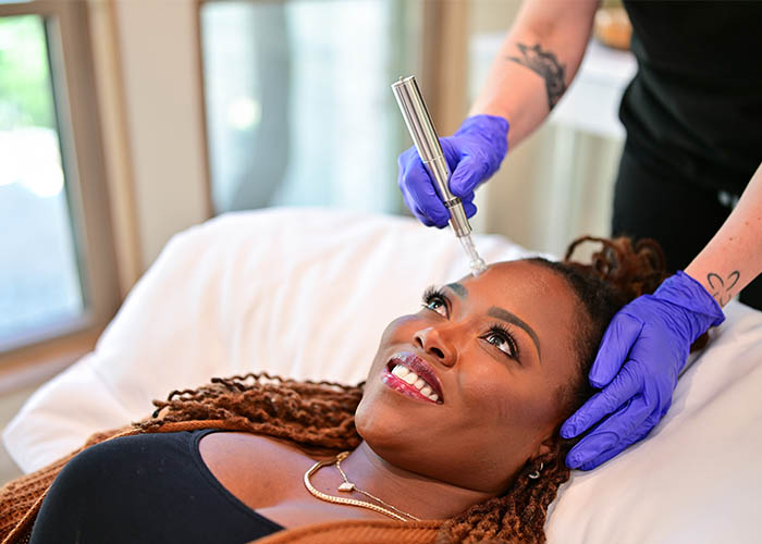 Aesthetician performing a facial treatment on a smiling woman lying down, using a device on her forehead. Saxon MD