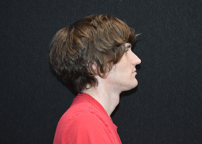 Profile view of a young man with shaggy brown hair, wearing a red shirt, against a black background. Saxon MD
