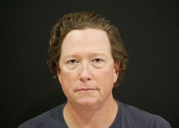 A middle-aged individual with short wavy hair against a black background, wearing a blue t-shirt and looking directly at the camera. Saxon MD