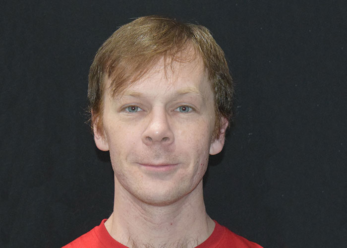 A man with light brown hair and a slight smile, wearing a red shirt, against a black background. Saxon MD