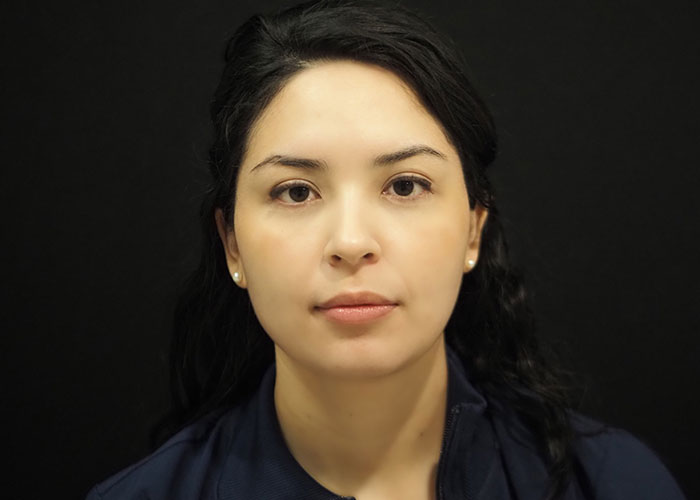 A woman with dark hair wearing a navy blue shirt against a black background. she has a neutral expression and pearl earrings. Saxon MD